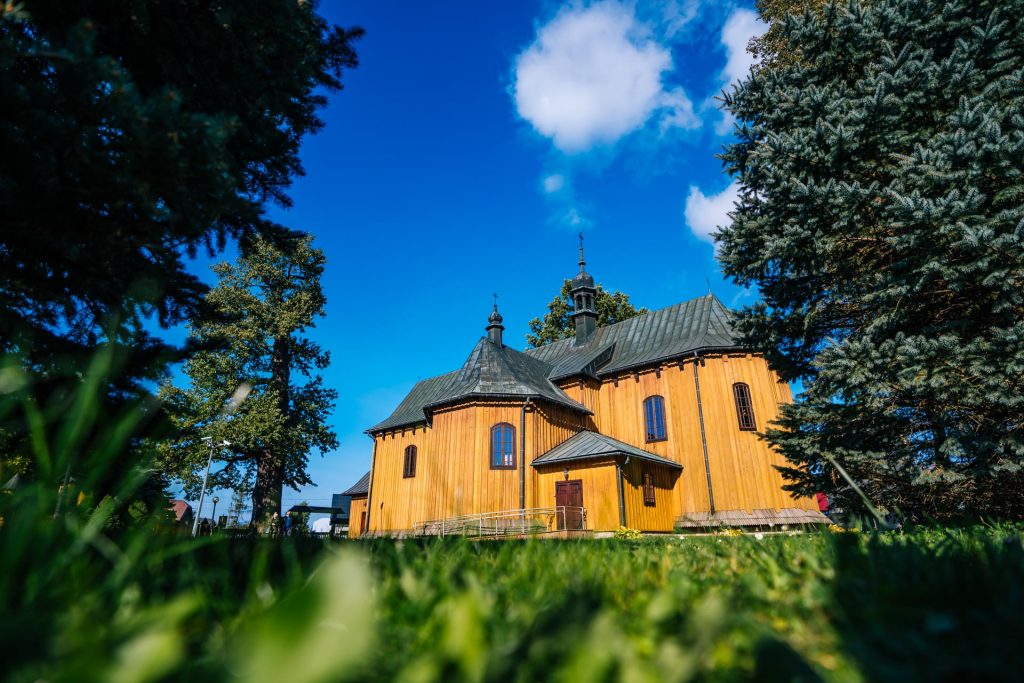 Drewniany kościół, dach blaszany. Przed kościołem liściaste drzewa.