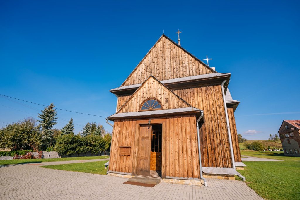Drewniany kościół, dach blaszany. Przed kościołem plac z kostki brukowej.