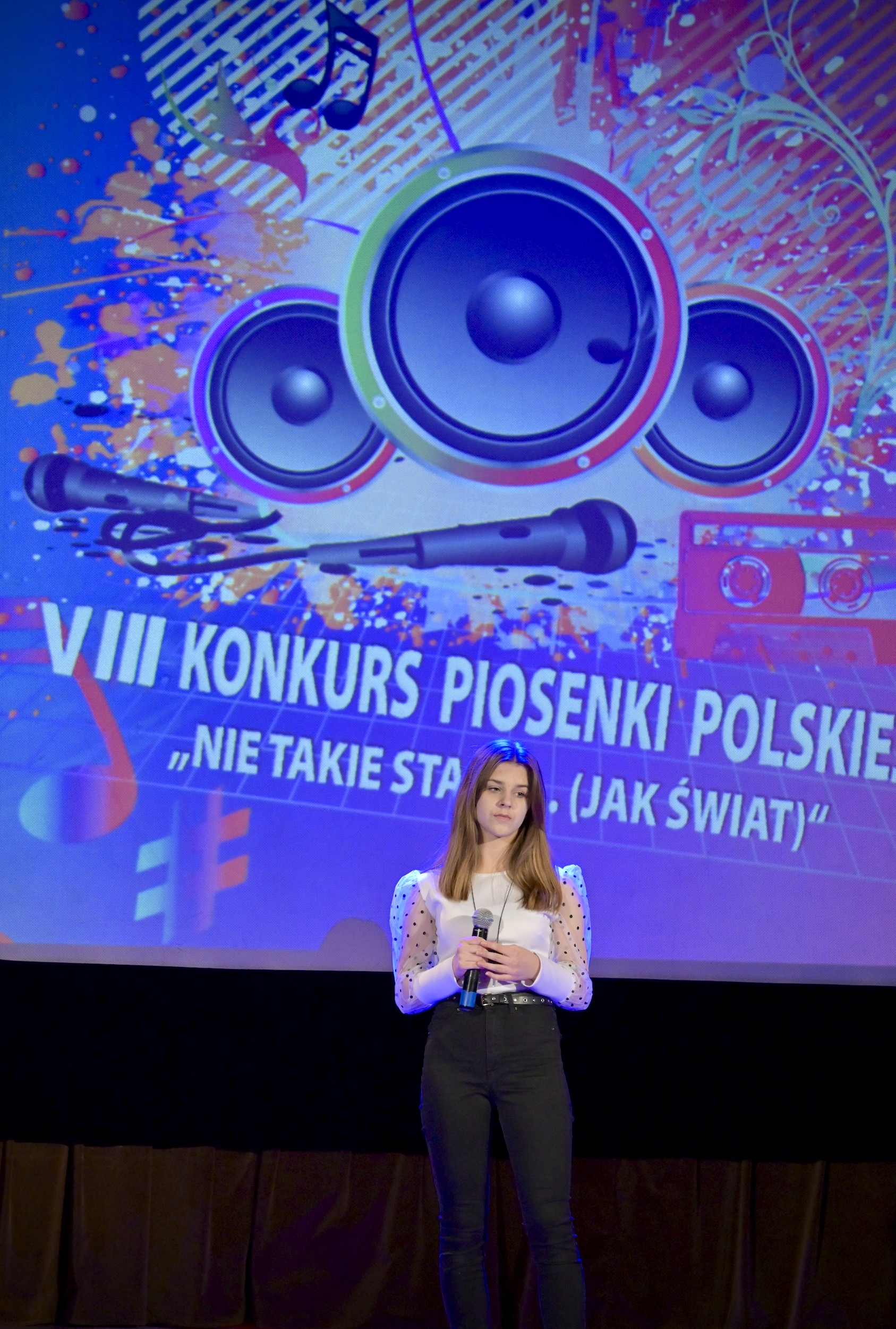 Dziewczyna z ciemnymi włosami, w białej bluzce i ciemnych spodniach śpiewa na scenie. W rękach trzyma mikrofon. Za nią na ekranie wyświetlone logo konkursu.