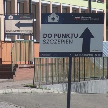 Tabliczka umoeszczona przed budynkiem z napisem do punktu szczepień. Za tabliczką schody do budynku z zieloną barierką. Obok drzewa.