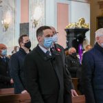 :Liczna grupa ludzi ubranych odświętnie ubranych stoi w ławkach w kościele. Na pierwszym planie stoi młody mężczyzna w garniturze i maseczce.