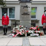 Dwie dziewczyny w czerwonych kurtkach stoją przy pomniku Józefa Piłsudskiego. Przed pomnikiem leżą wiązanki kwiatów. Pomnik stoi przed budynkiem urzędu.