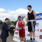 Młody mężczyzna wręcza dyplomy młodym zawodnikom . Za nimi baner z logami instytucjami wspierającymi.