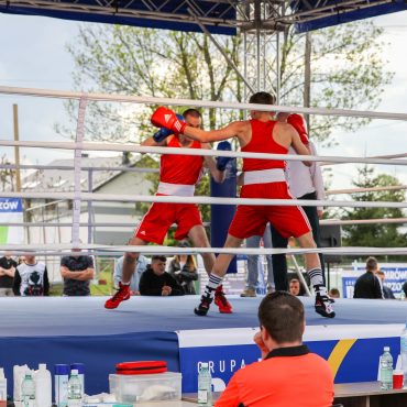 Na ringu dwóch zawodników w czerwonych strojach . Przed ringiem siedzi dwóch mężczyzn i obserwuje.