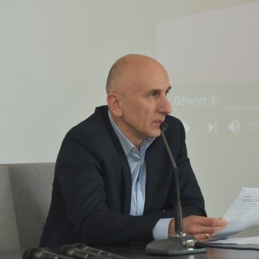 Starszy mężczyzna siedzi przy biurku i przemawia do mikrofonu. Za nim biała ściana.