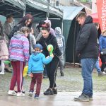 Dziewczyna w czarnej bluzie i spodniach oraz uszami myszka mini na głowie rozdaje dzieciom cukierki na rynku.