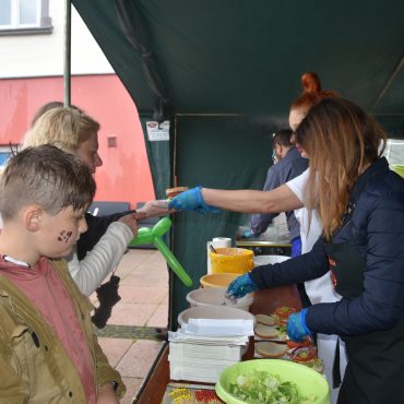 Dwie Panie pod namiotem serwują burgery. Przed kobietami stoją miski, naczynia, oraz jedzenie.