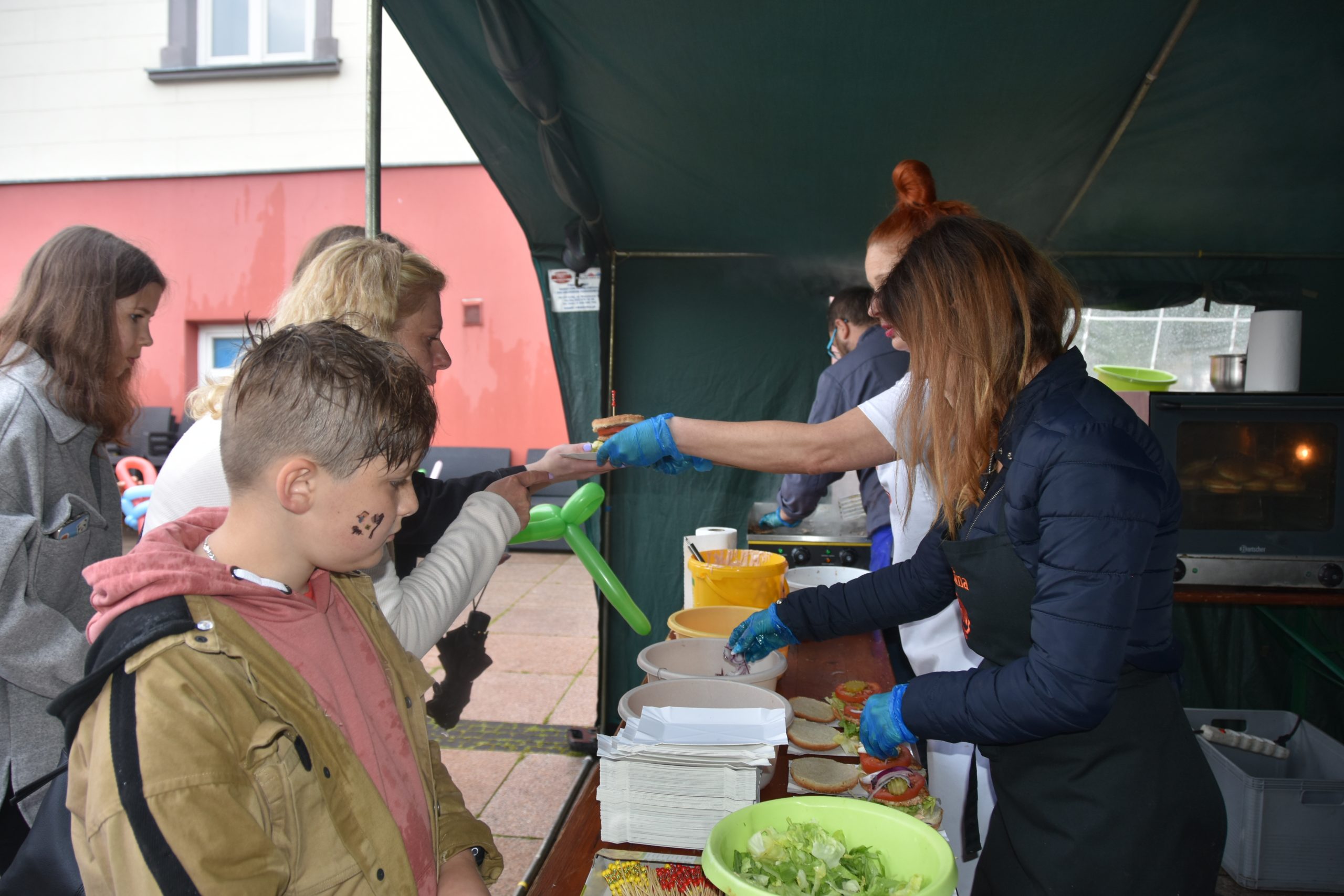 Dwie Panie pod namiotem serwują burgery. Przed kobietami stoją miski, naczynia, oraz jedzenie.