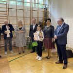 Mała dziewczynka w ciemnych spodanich i swetrze pozuje do zdjęcia z grupą starszych osób na sali gimnastycznej.