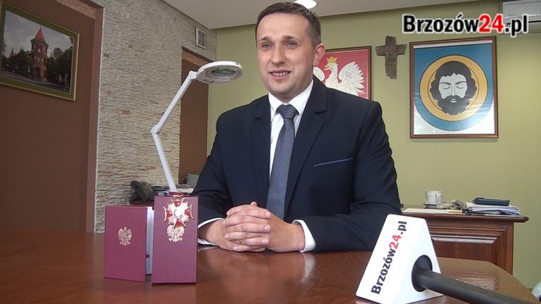 Młody mężczyzna w garniturze siedzi przy stoliku, przed nim leży legitymacja i odznaczenie.