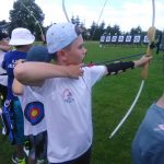 Młody chłopiec ubrany na sportowo w białą koszulkę i czapkę z daszkiem strzela z łuku.