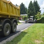 Żółta ciężarówka oraz rozścielacz do asfaltu stoją na drodze asfaltowej.