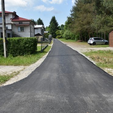 Nowa droga asfaltowa. Po prawej stronie zabudowa rodzinna, po prawej drzewa i krzaki.
