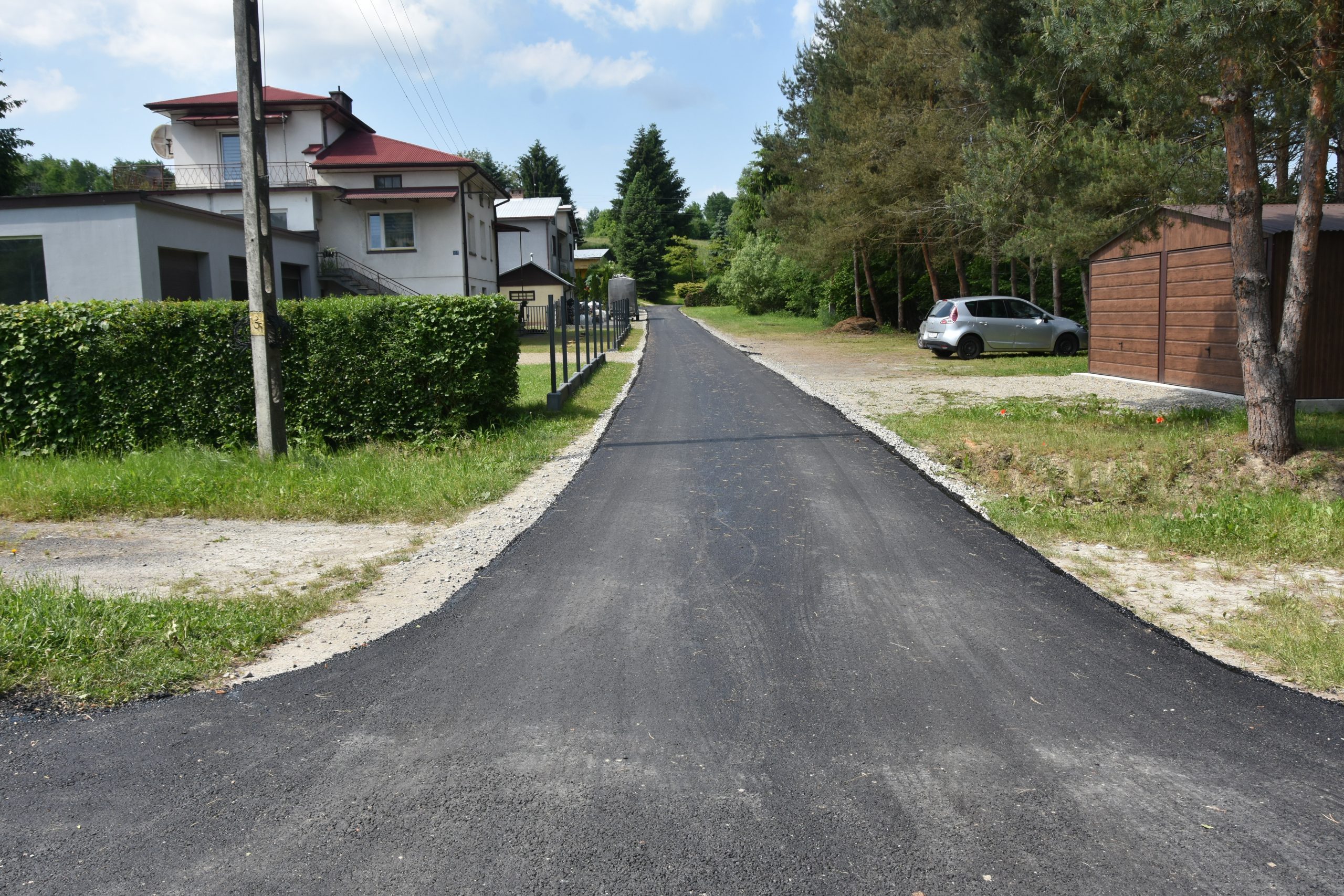 Nowa droga asfaltowa. Po prawej stronie zabudowa rodzinna, po prawej drzewa i krzaki.