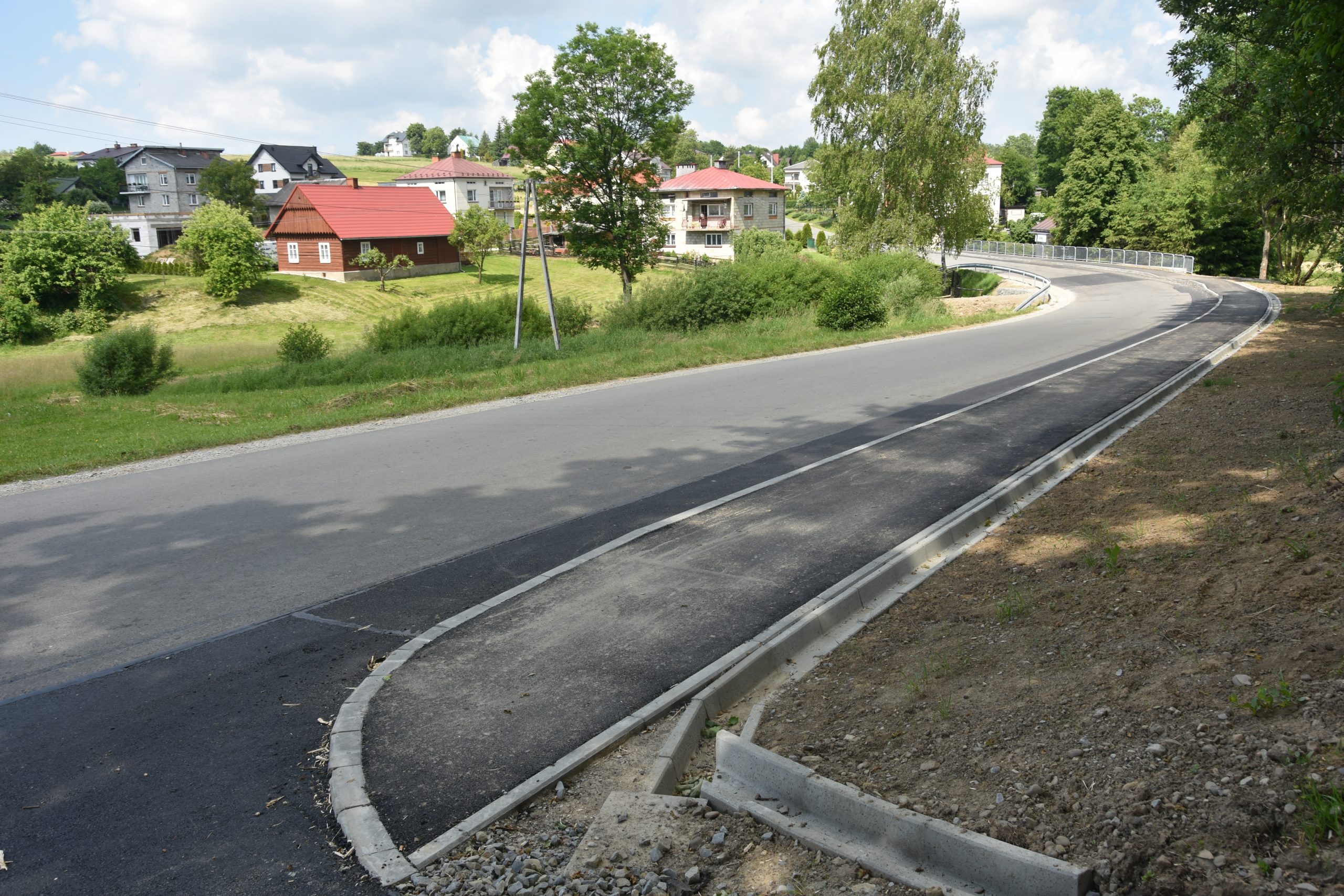 Nowy chodnik wzdłuż drogi asfaltowej. Po lewej stronie tereny zabudowane. .