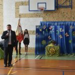 Mężczyzna w garniturze przemawia na sali gimnastycznej do dzieci. Za nim tablica z napisem wakacje oraz dzieci trzymające sztandar szkoły.