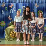 Dziewczynka stoi na sali gimnastycznej i trzyma w rękach mikrofon oraz teczkę. Za nią stoją dziewczynki w spódniczkach. Znajdują się one na sali gimnastycznej.