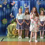 Dziewczynka stoi na sali gimnastycznej i trzyma w rękach mikrofon oraz teczkę. Za nią stoją dziewczynki w spódniczkach. Znajdują się one na sali gimnastycznej.