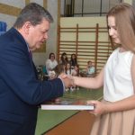 Mężczyzna w garniturze wręcza dziewczynce ubranej w białą koszulę i spodnicę na sali gimnastycznej książke.