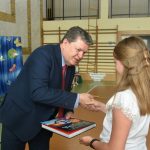 Mężczyzna w garniturze wręcza dziewczynce w białej koszuli na sali gimnastycznej książkę.