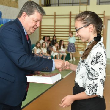 Mężczyzna w granatowym garniturze przekazuje dziewczynce książkę do ręki. Znajdują się oni na sali gimnastycznej w szkole.