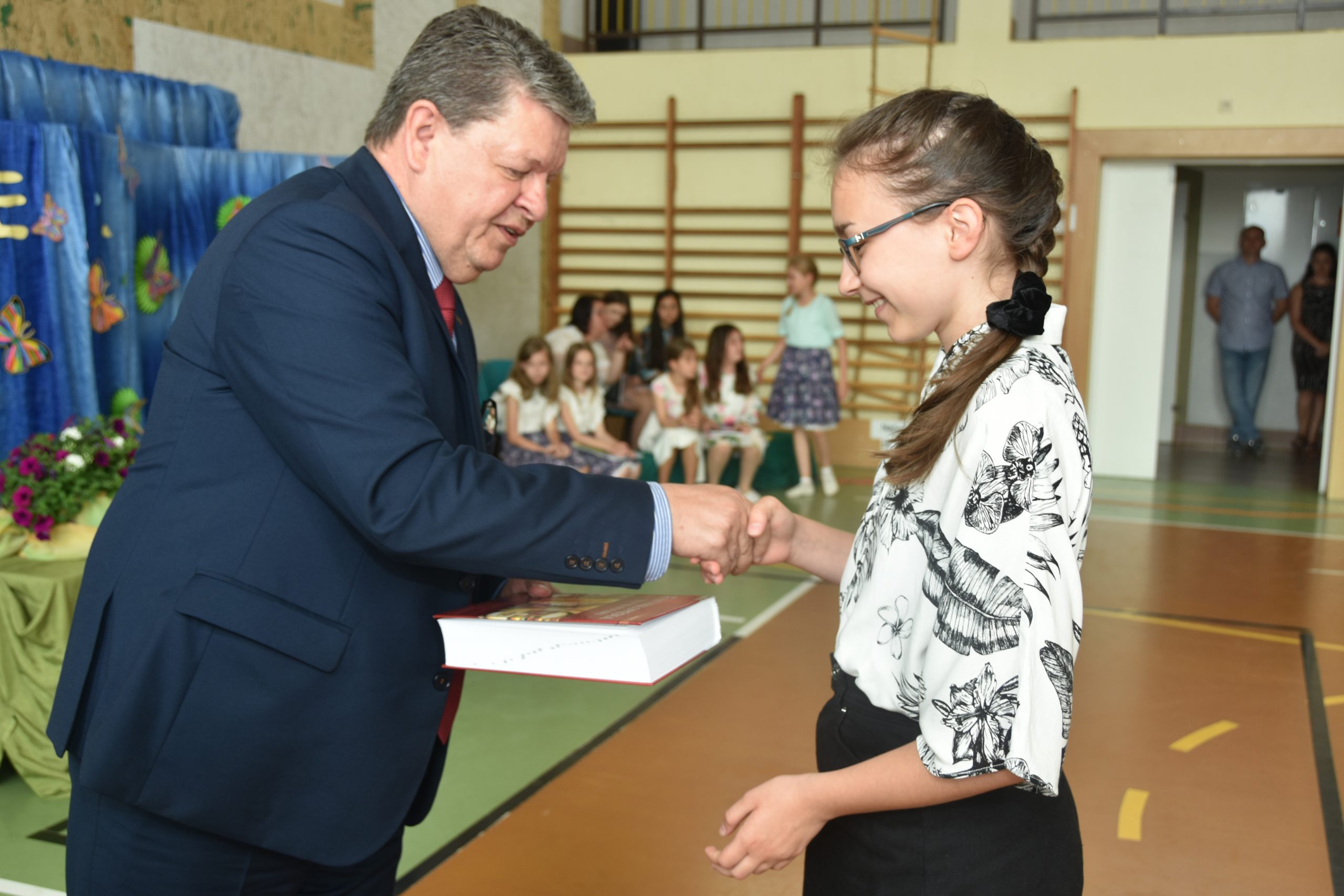 Mężczyzna w granatowym garniturze przekazuje dziewczynce książkę do ręki. Znajdują się oni na sali gimnastycznej w szkole.