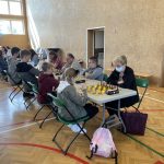 Dzieci i młodzież siedzą przy stolikach i grają w szachy na sali gimnastycznej.