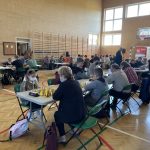 Dzieci siedzą przy stolikach i grają w szachy na sali gimnastycznej.