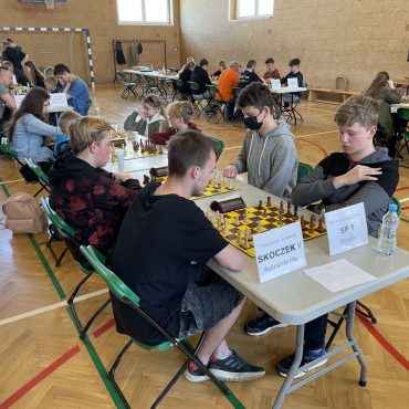 Dzieci siedzą przy stolikach i grają w szachy. Duża grupa dzieci znajduje się na sali gimnastycznej.