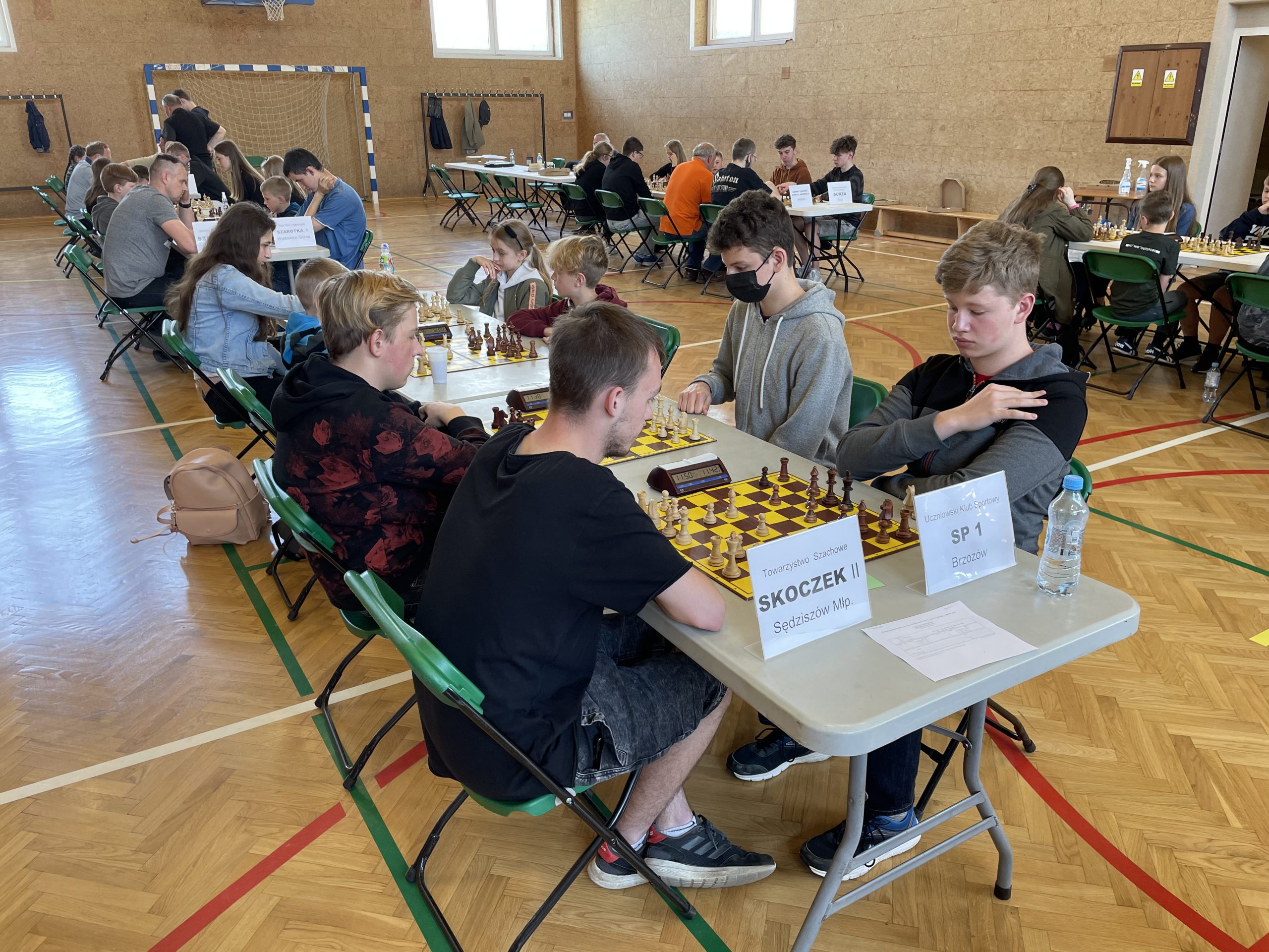 Dzieci siedzą przy stolikach i grają w szachy. Duża grupa dzieci znajduje się na sali gimnastycznej.