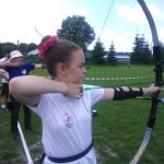 Dziewczynka ubrana na sportowo w białą koszulkę, na włosach gumka czerwona strzela z łuku.