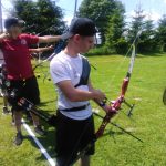 Młody chłopiec ubrany na sportowo w białą koszulkę i czapkę z daszkiem strzela z łuku.