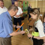 Starszy mężczyzna w niebieskiej koszuli wręcza dziewczynce z warkoczykami drobne gadźety za udział w turnieju.