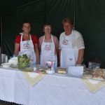 Trzy starsze panie ubrane w fartuszki stoją przy stole, na którym serwowane są różnego rodzaje potrawy.