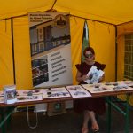 Pod namiotem żółtym stoisko z praca plastycznymi, na ławce siedzi młoda dziewczyna i trzyma pracę w rękach.