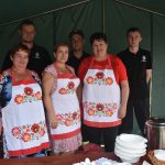 Trzy panie w fartuchach i trzech strażków ochotników stoją przy stole pod namiotem, w którym serwowane są potrawy.