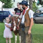 Jedna dziewczynka siedzi na kucyku, druga stoi przy niej. Obok nich stoi mężczyzna ubrany w strój kowbojski i trzyma konia.