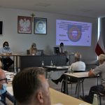 Grupa osób siedzi przy stolikach w sali konferencyjnej. Na ścianie wyświetlane sa informacje z projektora.