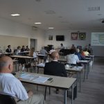 Grupa osób - radnych siedzi na sali konferencyjnej przy stolikach. Przed nimi na ścia nie wyświetlane są informacje z projektora.