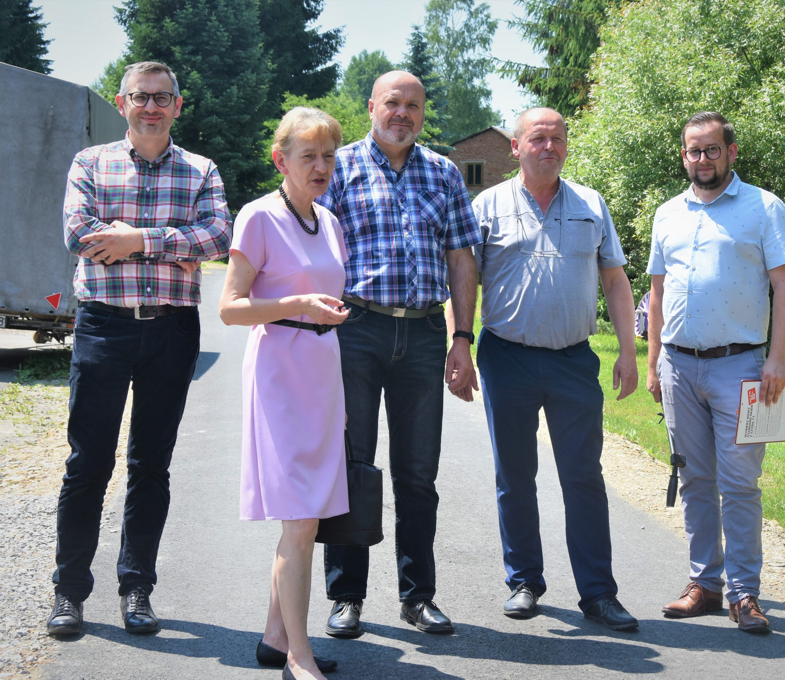 Grupa osób pozujących do zdjęcia. W tle asfaltowa droga i drzewa.