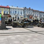 Stare wojskowe samochody zaparkowane na rynku. Obok nich przechadzają się osoby w różnym wieku. W tle kamienice.