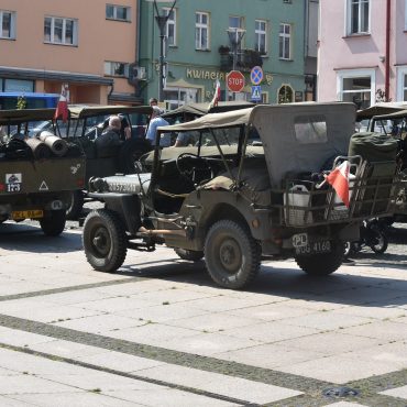 Stare wojskowe samochody na rynku miejskim. W tle zabudowa.