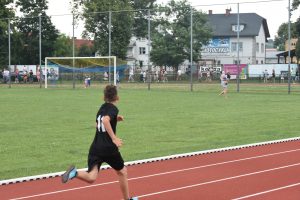 Chłopak biegnący po boisku. W tle osoby w różnym wieku.