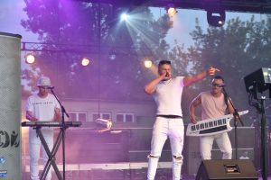 Trzech mężczyzn ubranych na biało, prezentujący występ muzyczny na scenie.