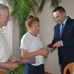 Mężczyzna w garniturze przekazuje wyróżnienie starszemu mężczyźnie i starszej kobiecie.