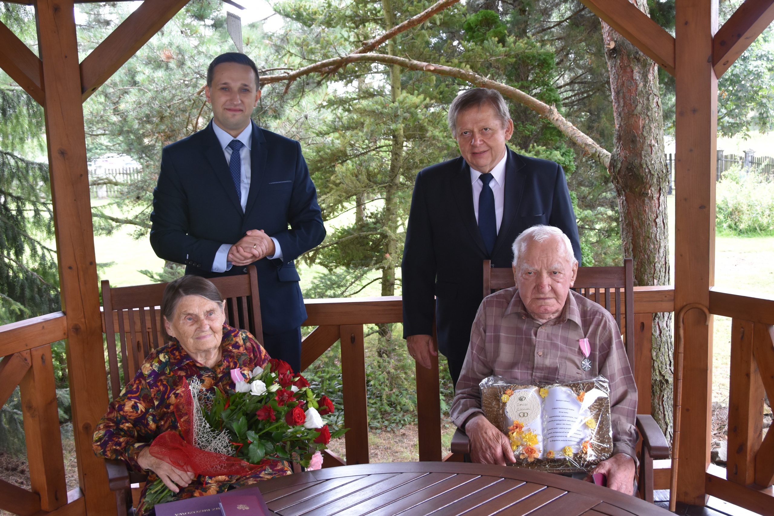 Dwóch stojacych meżczyzn w garniutrów i para staruszków siedząca przy stole.