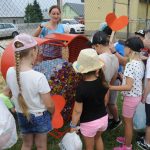 Dzieci pod nadzorem opiekunów wrzucają nakrętki do metalowego serca. W tle parking i samochody osobowe.