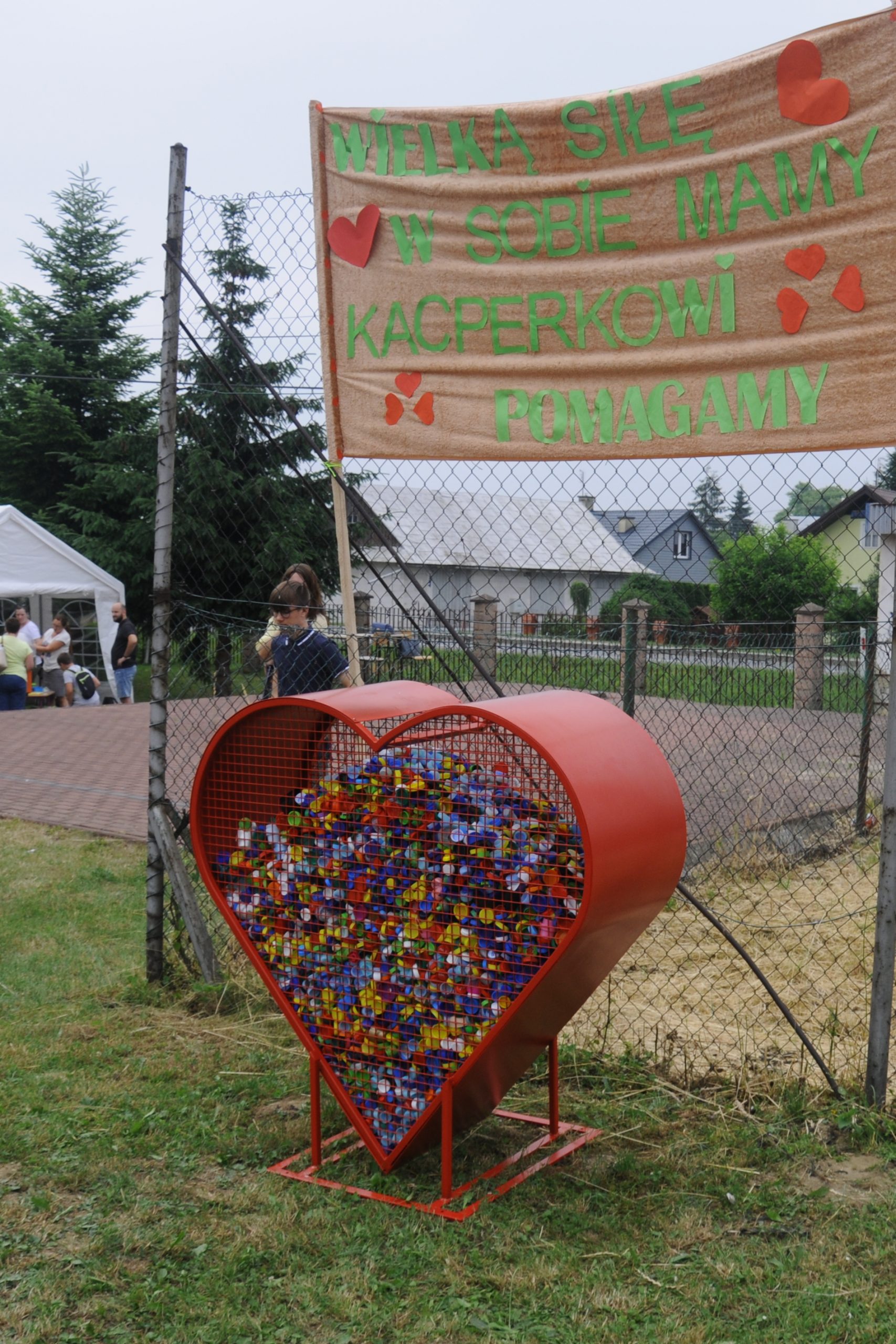 Czerwone, metalowe serce wypełnione zakrętkami. Za nim znajdują się uczestnicy festynu.