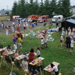 Dzieci, młodzież i dorośli zebrani na festynie przy Szkole Podstawowej w Humniskach. W tle widać samochody, m.in. strażackie i policyjne.