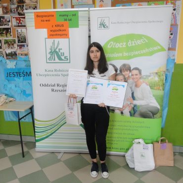 Dziewczyna w czarnych spodniach i białej bluzce stoi przed tablicami promującymi KRUS.